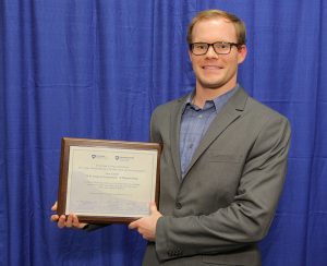 At the 2016 Penn State College of Medicine Innovation Awards, pharmacology department PhD student Sam Linton was recognized for his service to support commercialization and the research enterprise at the College of Medicine and Hershey Medical Center. Linton is pictured in front of a photo backdrop, smiling and holding a plaque with his award.