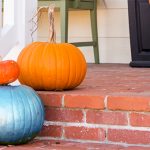 A close-up of a brick front porch with a white railing on one side. Next to the railing are three pumpkins - two orange and one blue.