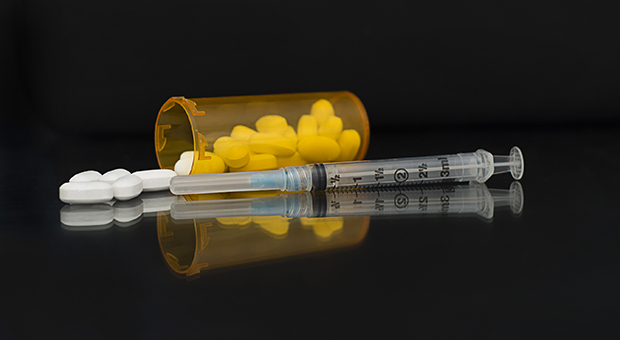 A syringe is pictured with an open bottle of prescription medicine behind it on its side with pills spilling out. Both are in front of a black background.