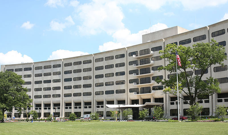 The Penn State College of Medicine Crescent facade is seen at 500 University Drive in Hershey, PA, framed by grass below and a blue sky with clouds above. In front of the building to the right, two flagpoles can be seen displaying the American and Pennsylvania flags.