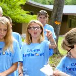 A teenage girl extends her fingers to see if an orange butterfly will alight upon them as a group of other teens stands around her, outdoors, all wearing blue Camp Lionheart T-shirts.
