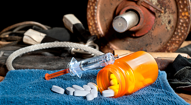 A syringe sitting atop a spilled bottle of pills on top of a blue rag, with a rusted weightlifting plate attached to a bar sitting next to jump ropes in the background.