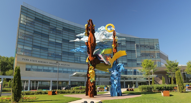The exterior of the Penn State Health Children's Hospital is shown in the background, with a large abstract sculpture in focus in the foreground.