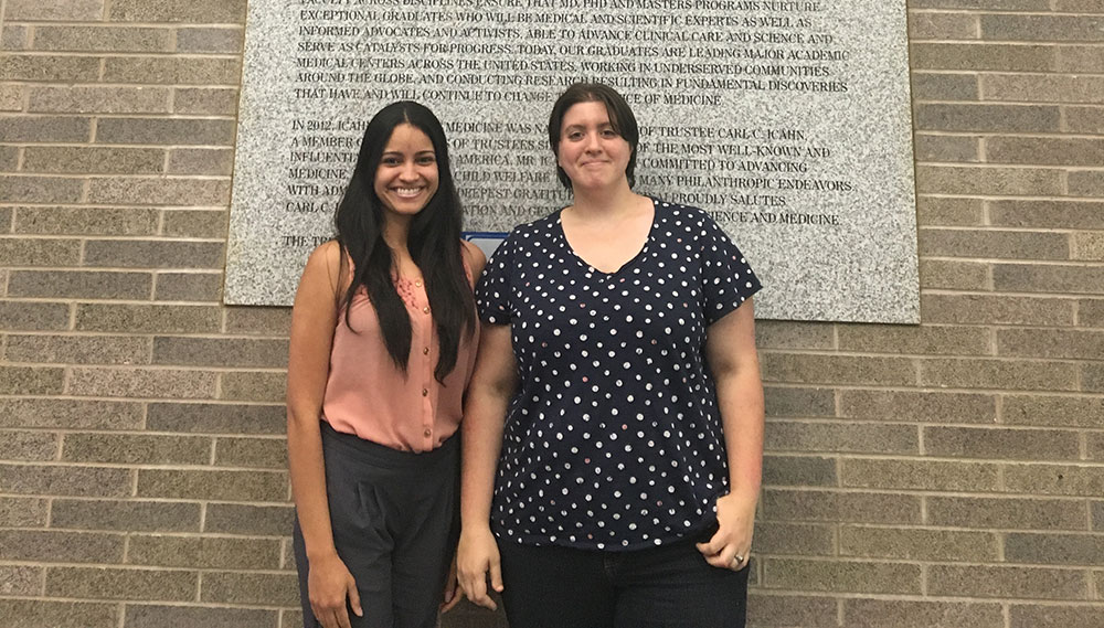 Nathalie Fuentes Ortiz, left, and Jesica James are pictured at Icahn School of Medicine at Mount Sinai in New York City. The two women are standing in front of a brick wall and the bottom part of a large plaque describing the school can be seen behind them from shoulder-height and up.