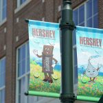 Two banners depicting cartoon candy characters are seen on a light pole in downtown Hershey, PA, in summer 2016, with a brick building with large glass windows in the background.