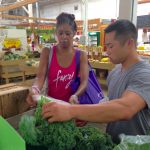 John Chan, a medical student who helped develop the Prevention Produce program, helps a participant choose vegetables in 2015. Chan is pictured at right, holding large, leafy green vegetables. A female participant in the program is seen at left, looking at the produce with a purple tote bag over her shoulder.