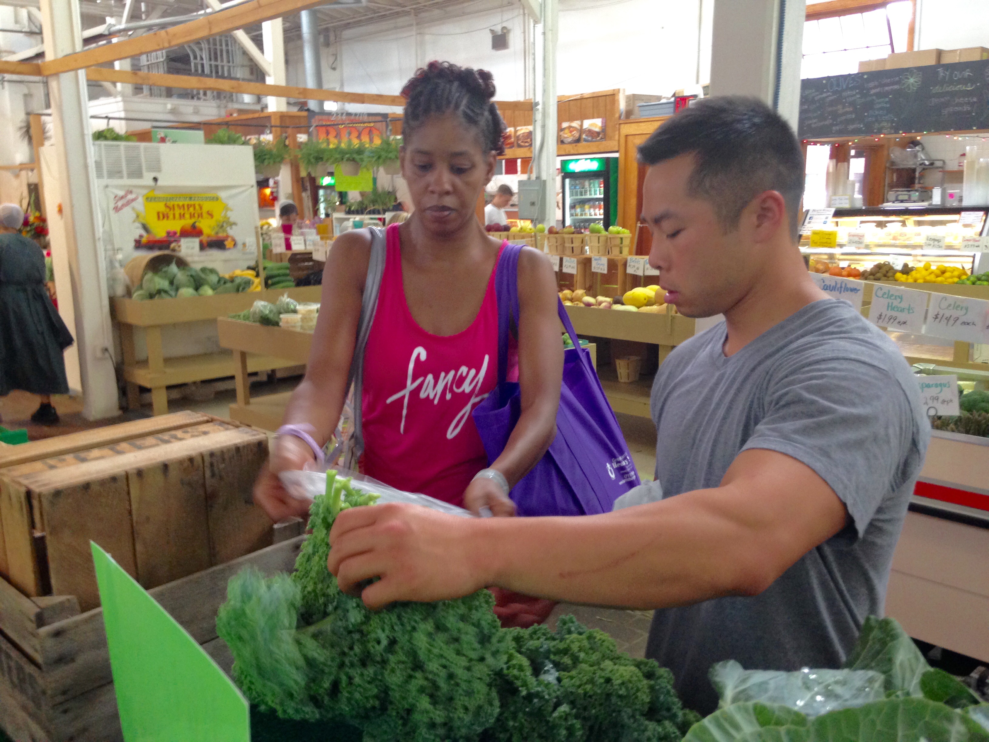 John Chan, a medical student who helped develop the Prevention Produce program, helps a participant choose vegetables in 2015. Chan is pictured at right, holding large, leafy green vegetables. A female participant in the program is seen at left, looking at the produce with a purple tote bag over her shoulder.