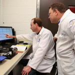 Todd Umstead, Research Project Manager, left, and E. Scott Halstead, MD, PhD, Director, right, are seen at work in the Pulmonary Immunology & Physiology Core in 2015. Umstead is at the center of the photo, looking left toward a computer screen and framed by lab equipment on the table in front of him. Umstead looks over his shoulder from the right of the photo.