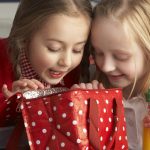 Two girls look excitedly into a reusable lunch bag (red, with white polka dots) as the one clutches orange juice.
