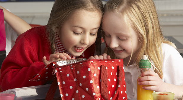 Two girls look excitedly into a reusable lunch bag (red, with white polka dots) as the one clutches orange juice.