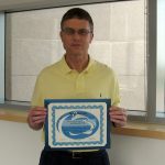 Scot Kimball was voted by the graduate students of Penn State College of Medicine as the best overall professor of the 2014-15 academic year. Kimball is pictured standing in front of a window in a yellow polo shirt, holding a certificate noting his honor.