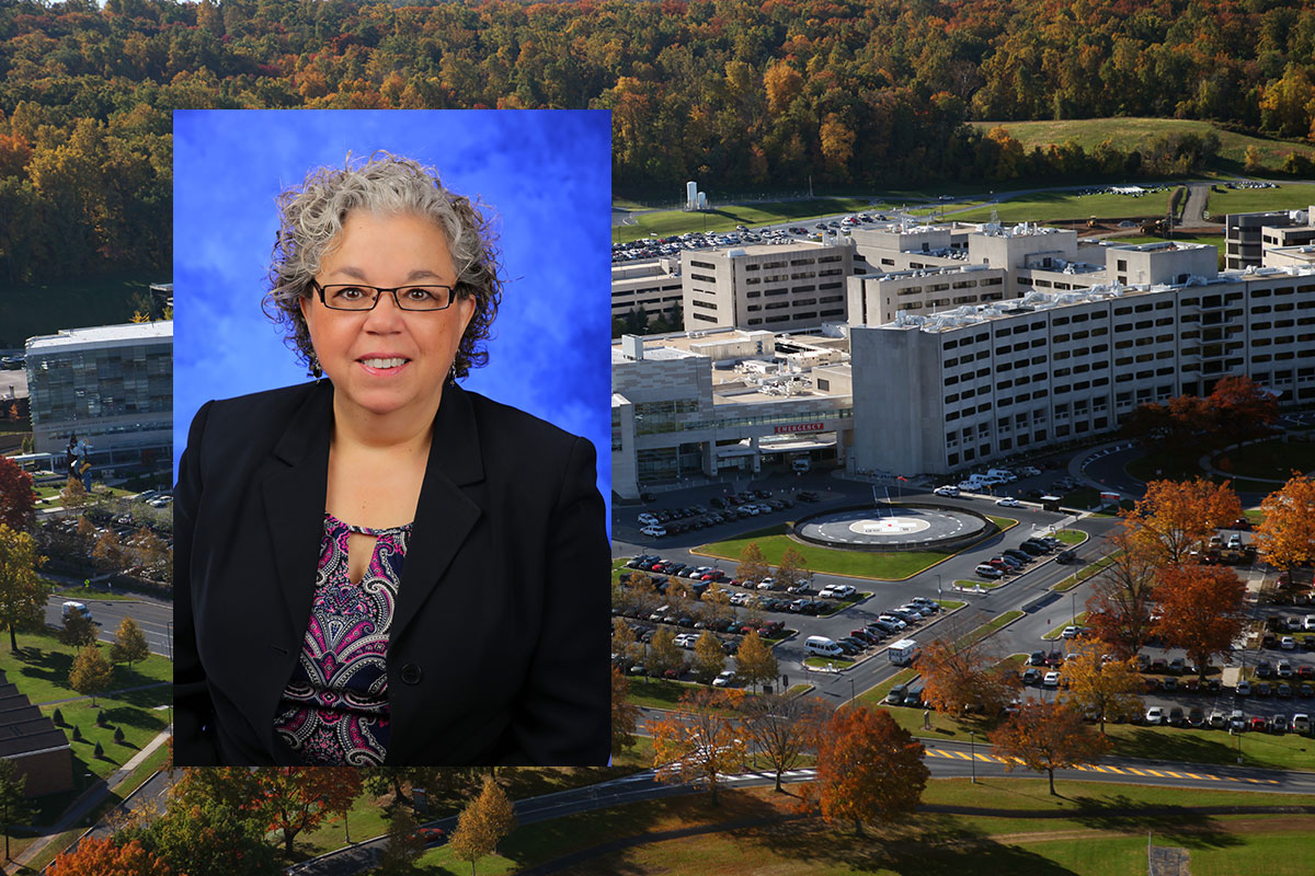 Barbara Ostrov, MD, was named associate dean for faculty and professional development at Penn State College of Medicine in September 2016. Ostrov is pictured wearing a purple patterned blouse, dark blazer and glasses, in front of a blue photo background. Her photo is superimposed on the left side of an aerial image of Penn State College of Medicine's Hershey campus, with the College Crescent visible at right.