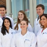 Six new anatomy students in white coats stand in front of an entryway.