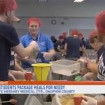 A number of physician assistant students are shown, wearing hairnets and matching blue shirts, around a table packaging food into containers to be sent around the world.