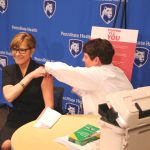 A nurse gives PA Health Secretary Karen Murphy a flu shot in her left arm. Both are seated at a small table, with a 'Penn State Health' backdrop in the background.