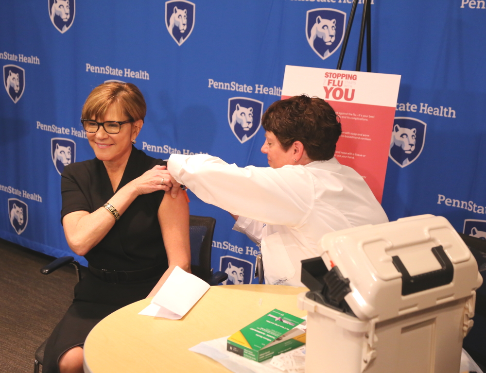 A nurse gives PA Health Secretary Karen Murphy a flu shot in her left arm. Both are seated at a small table, with a 'Penn State Health' backdrop in the background.