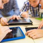 A close-up of four children huddled around a table sharing three tablet computers.