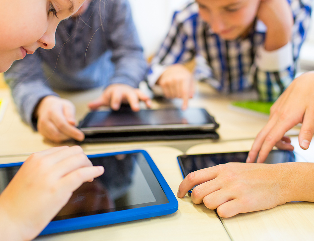 A close-up of four children huddled around a table sharing three tablet computers.