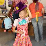 A young girl tips her purple hat, while wearing a dress-like costume with a floral print.