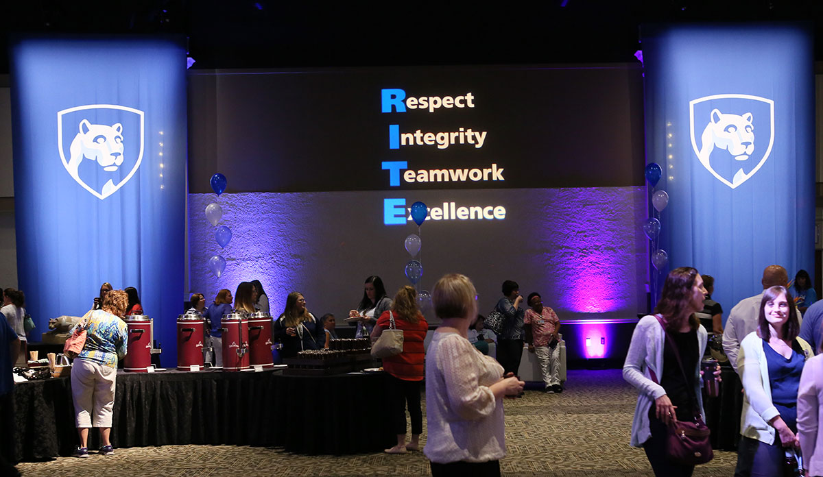 At each year's Inspired Together assembly, faculty, students and staff are honored for exemplifying the organization's RITE values of respect, integrity, teamwork and excellence. The 2016 awards assembly is pictured, with the RITE values projected onto a blue screen between the images of two Nittany Lion shields. People attending the assembly are seen in a crowd in front of the screen.
