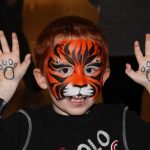 A child in tiger face paint holds his hands up, showing a paw print tattoo in each hand.