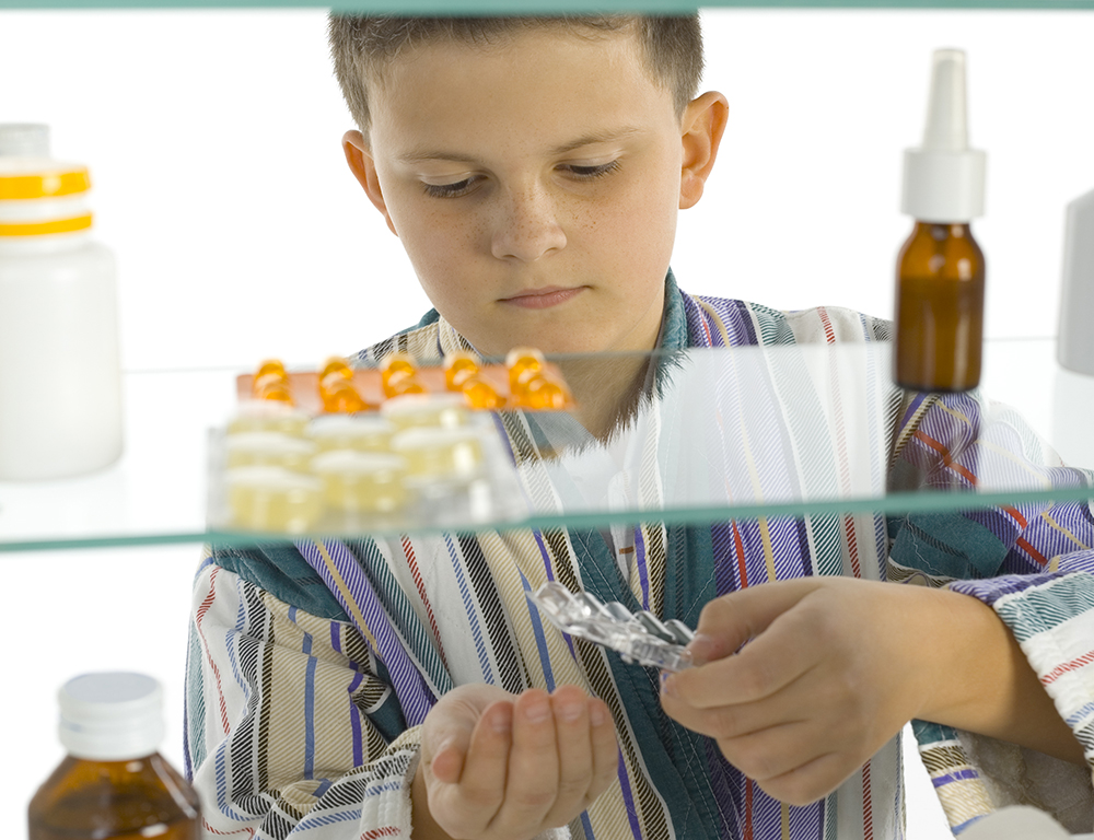 A child is seen through a medicine cabinet punching pills out into his hand. In the foreground, the cabinet contains several different medications.