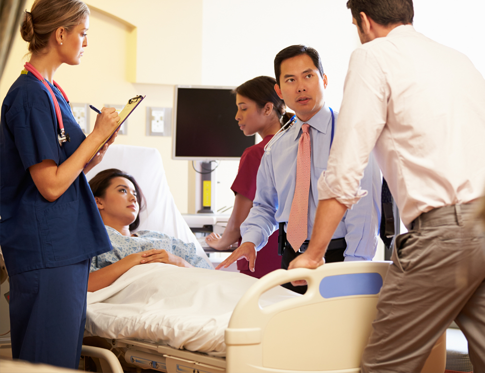 Physicians and nurses stand around the bed of a female patient, talking to one another.