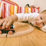A child lays on the floor while playing with a toy train set.