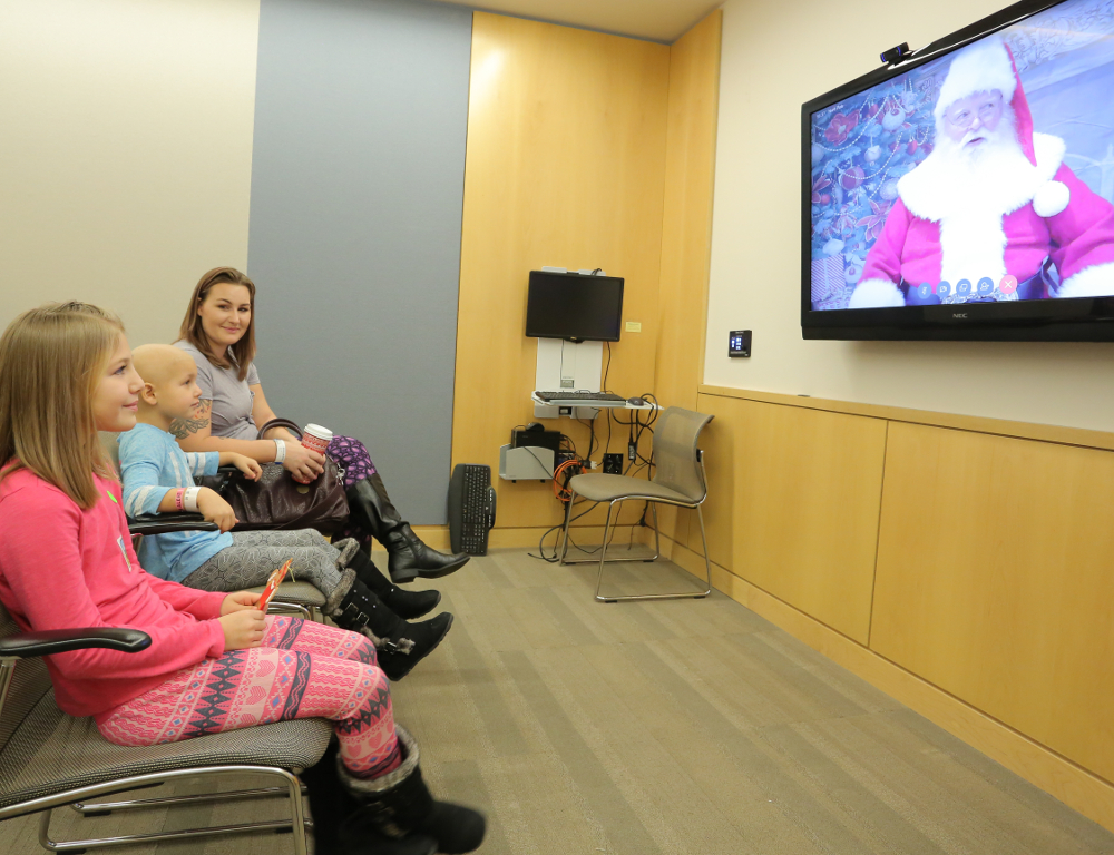 Three people -- two children and an adult -- are seated at left, viewing a screen at right that displays Santa Claus.
