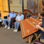 Two musicians, playing a hammer dulcimer and harp, perform as three people sitting nearby listen.