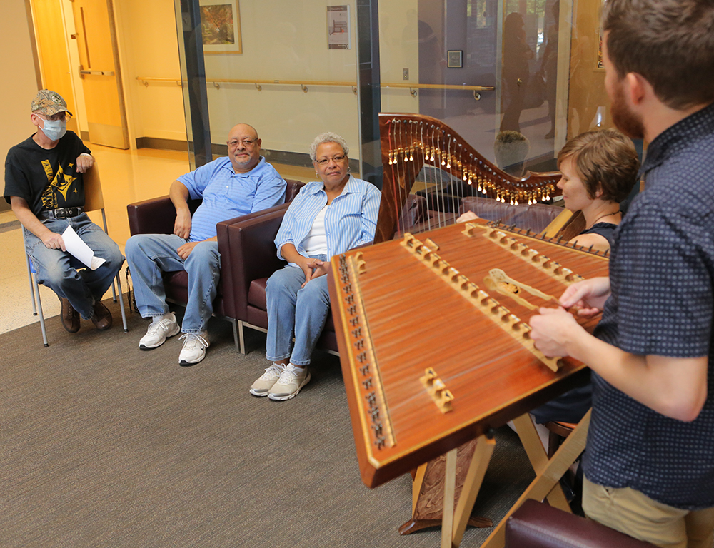 Two musicians, playing a hammer dulcimer and harp, perform as three people sitting nearby listen.