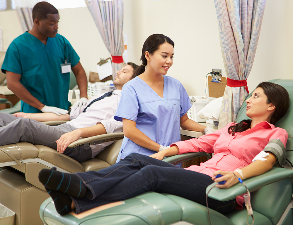 In the foreground, a female nurse tends to a female patient who™s donating blood. In the background, a male nurse tends to a male patient who™s donating blood.