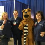 Three nurses and the Nittany Lion pose for a photo, in front of a blue pipe-and-drape background.