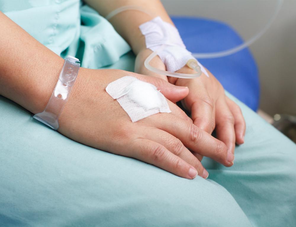 A close-up of a hospital patient™s hands, partially folded in their lap. One hand is connected to an IV line, the other has a white bandage on it.
