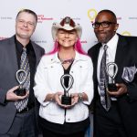 Nipsey, Jen Shade and Earl David Reed pose for a photo while holding small trophies, standing in front of a backdrop that has several CMN Hospitals logos.