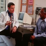 A physician sits at his desk talking with a patient. The physician is wearing a white coat and a stethoscope; the patient is wearing a blue shirt and dark pants.