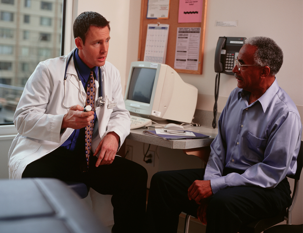 A physician sits at his desk talking with a patient. The physician is wearing a white coat and a stethoscope; the patient is wearing a blue shirt and dark pants.