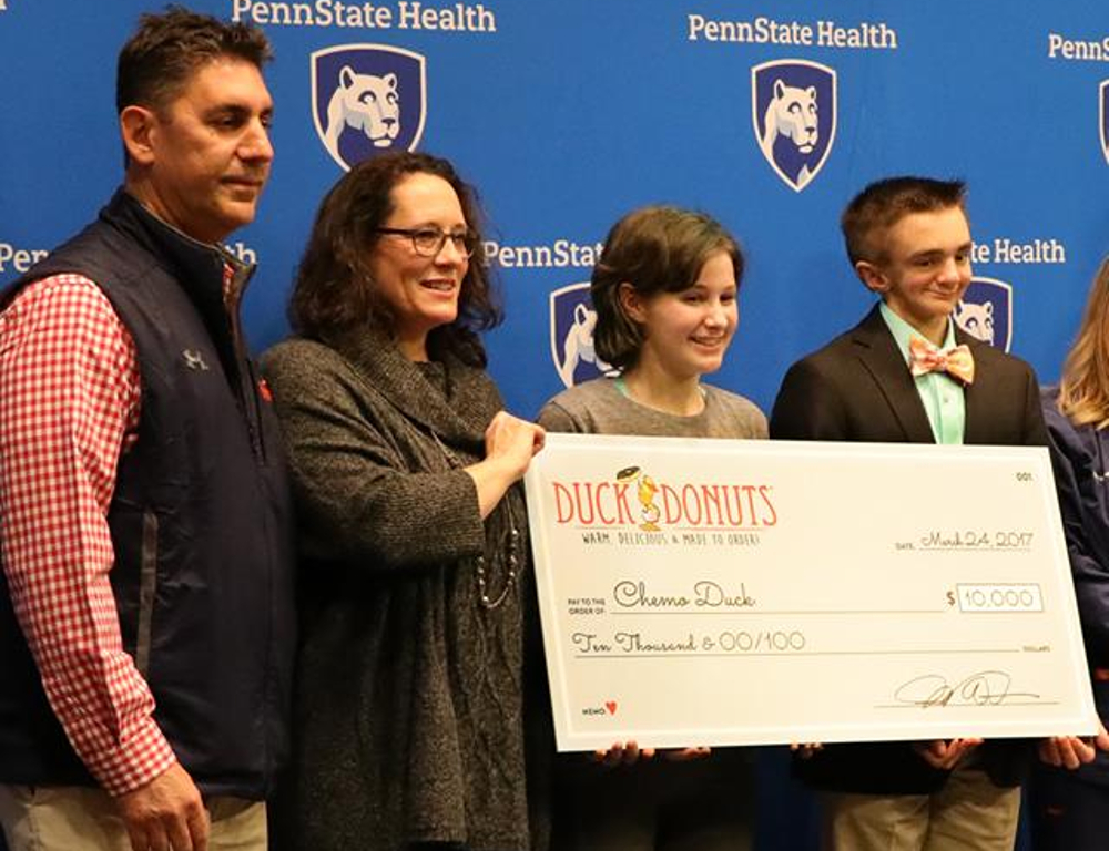 Four people pose holding an oversized check. They stand in front of a blue backdrop with the Penn State Health logo.