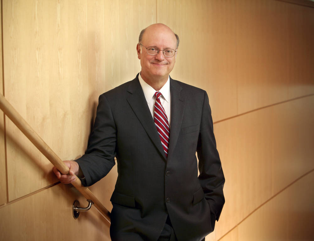 Dr. A. Craig Hillemeier poses in a dark suit with a red tie. His right hand is resting on a staircase bannister. The background is a wooden wall.