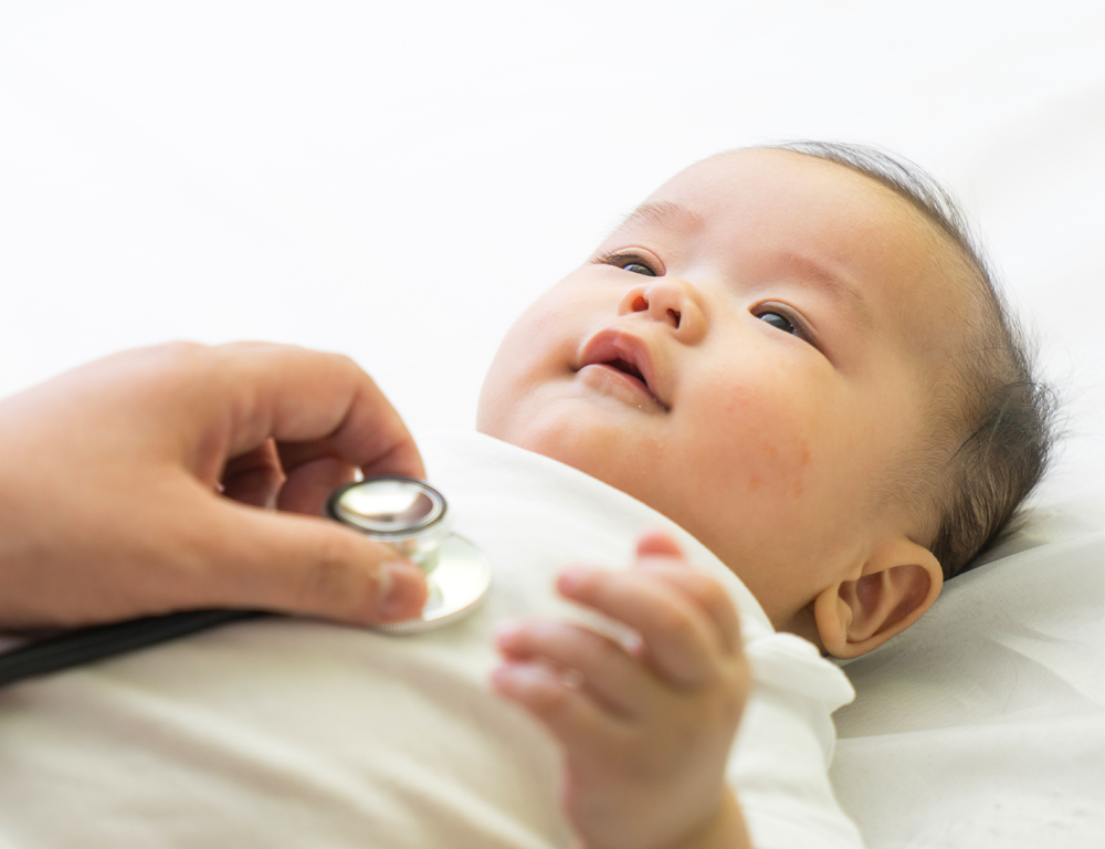 An infant wearing white lays on a white sheet. An adult is holding a stethoscope on the infant™s chest.
