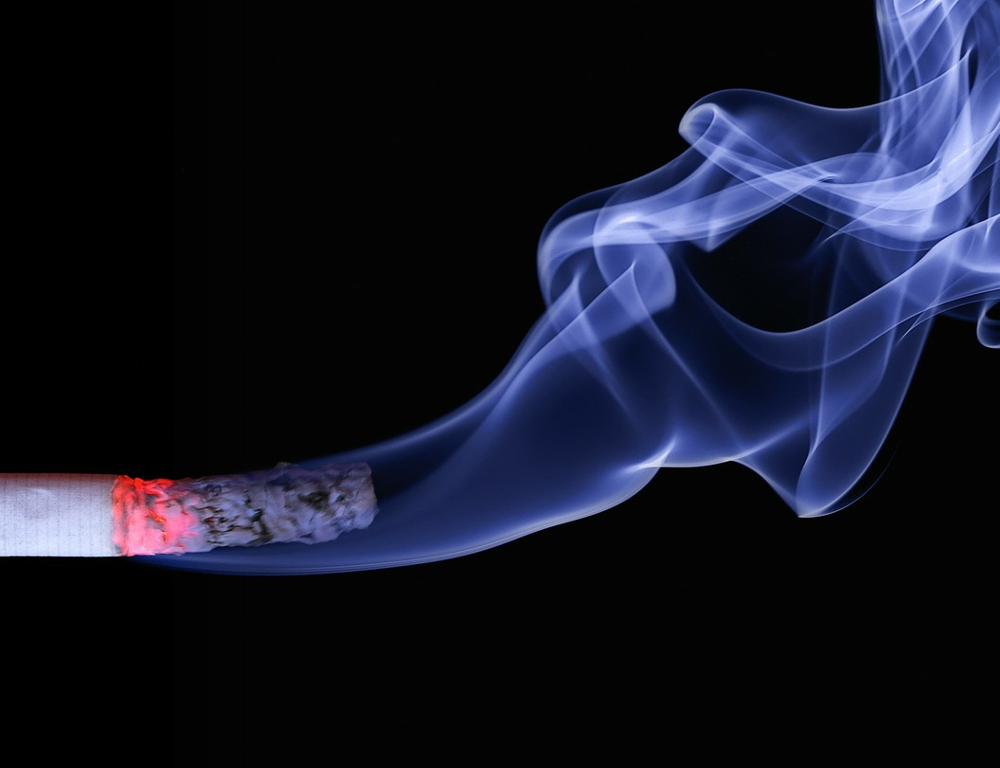 Close-up of a lit cigarette with a red tip and some residual ash on the end, with white smoke wafting upward, all against a black background.