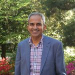 Dr. Krish Sathian poses for a photo wearing a patterned shirt and blue jacket. Trees are in the background.