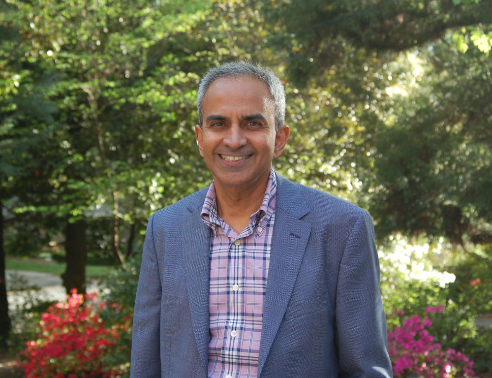 Dr. Krish Sathian poses for a photo wearing a patterned shirt and blue jacket. Trees are in the background.