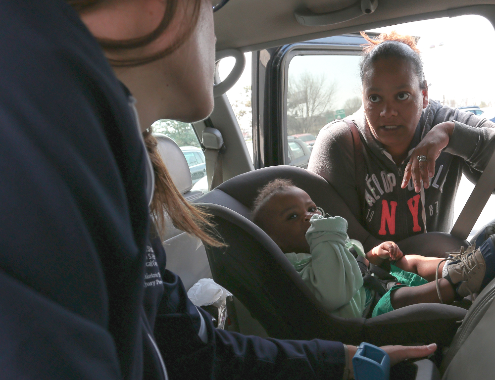 Two women speak to each other across the back seat of a car. An infant in a child safety seat is between them.