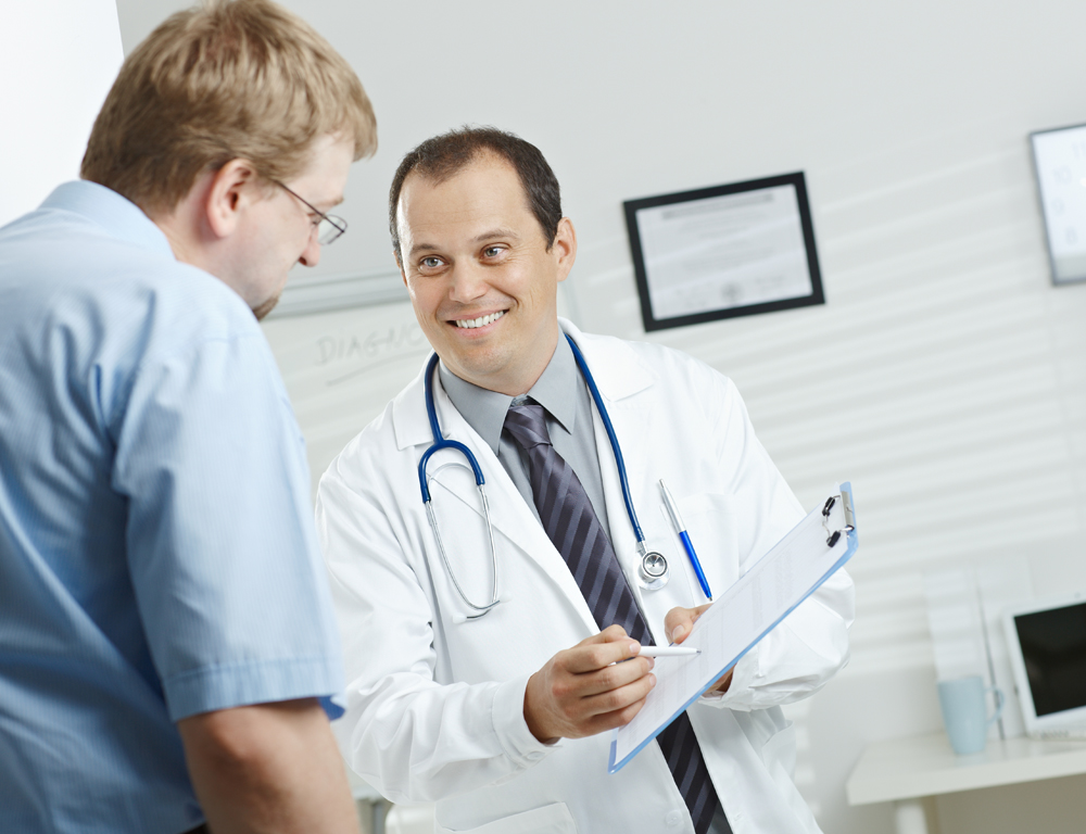 A male physician wearing a white coat and a stethoscope around his neck holds a clipboard with his left hand, using a pen in his right hand to point to the clipboard as a male patient looks down at it.