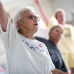 An older woman in the foreground is exercising by raising her right arm in the air. Two other people in the background are slightly out of focus.