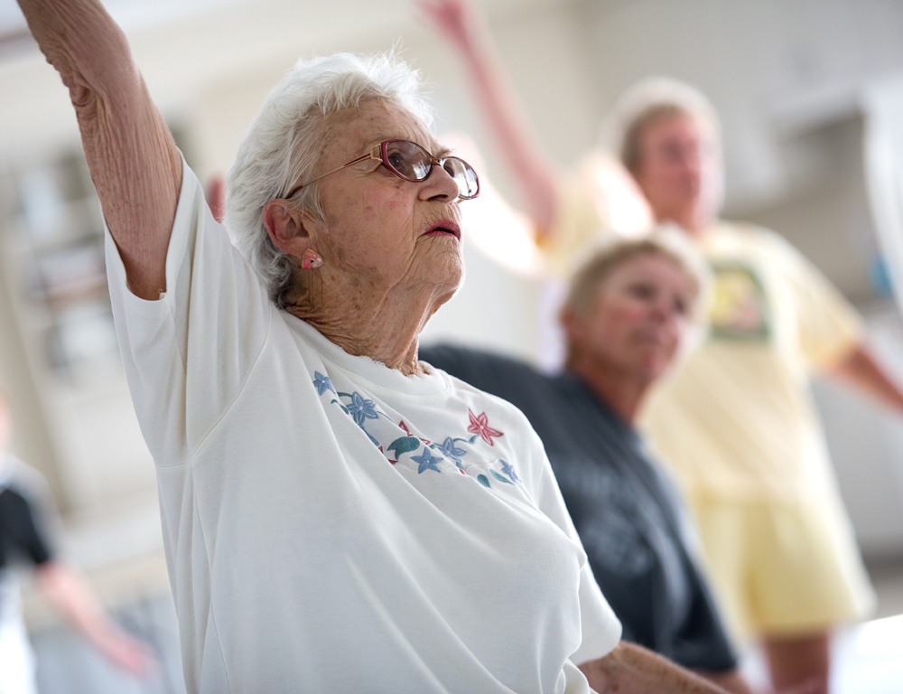 An older woman in the foreground is exercising by raising her right arm in the air. Two other people in the background are slightly out of focus.