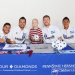 Four City Islanders soccer players sit at a table, surrounding a young child holding a soccer ball. The table has a blue tablecloth with the Four Diamonds and Penn State Health Children's Hospital logos.
