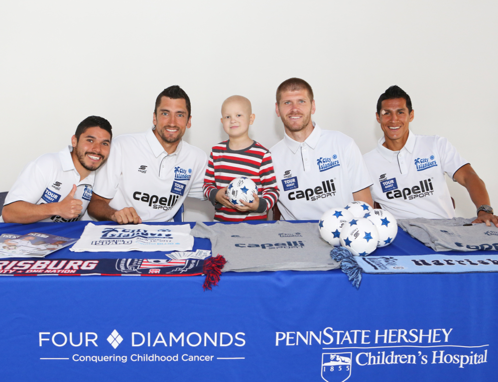 Four City Islanders soccer players sit at a table, surrounding a young child holding a soccer ball. The table has a blue tablecloth with the Four Diamonds and Penn State Health Children's Hospital logos.