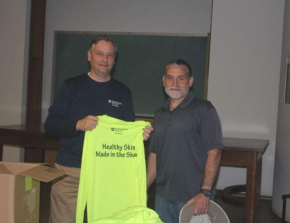 Two men pose for a photo. The man on the left holds a neon yellow shirt with the words Health Skin Made in the Shade printed on it. A table and chalkboard are in the background.
